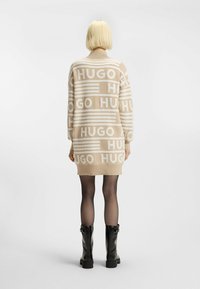 Knit-kjole i beige med hvit "HUGO"-bokstaver, lange ermer og ribbestrikkede mansjetter. Kombinert med svarte ankel støvler. Glatt tekstur.