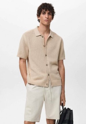 Camicia beige a maniche corte in maglia con un motivo testurizzato, chiusura con bottoni e colletto, abbinata a pantaloni corti chiari e una borsa nera.