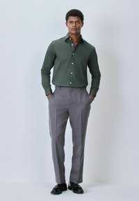 Grünes langärmliges Hemd mit Knöpfen, kombiniert mit grauen maßgeschneiderten Hosen und schwarzen Schuhen. Das Hemd bietet eine klassische Passform und hat eine glatte Stofftextur.