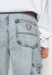 Ljust tvättade denimjeans med en triangel-logotyp "GUESS" på bakfickan. Har sömnadsdetaljer och en texturerad yta.