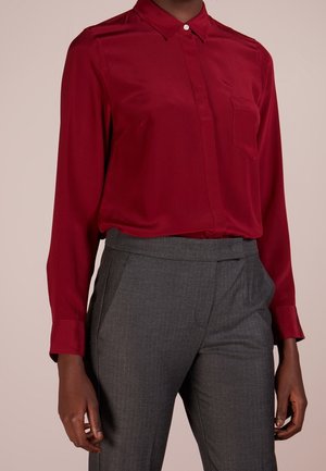 Osoba ubrana w czerwoną, długorękawową koszulę zapinaną na guziki, włożoną w wysokowalne, szare spodnie o kroju na miarę, na jednolitym tle.