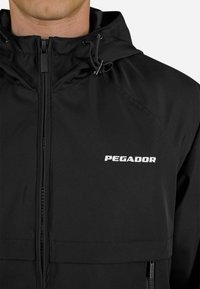 Čierna kapucňová bunda s predným zipsom, nastaviteľnou šnúrkou a bielym logom "PEGADOR" na hrudi, nosená osobou od krku po pás.