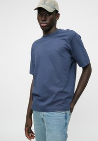 Μπλέ βαμβακερό t-shirt με κανονικό λαιμό, κοντό μανίκι και χαλαρή εφαρμογή, συνδυασμένο με ανοιχτόχρωμο τζιν και μπεζ καπέλο.