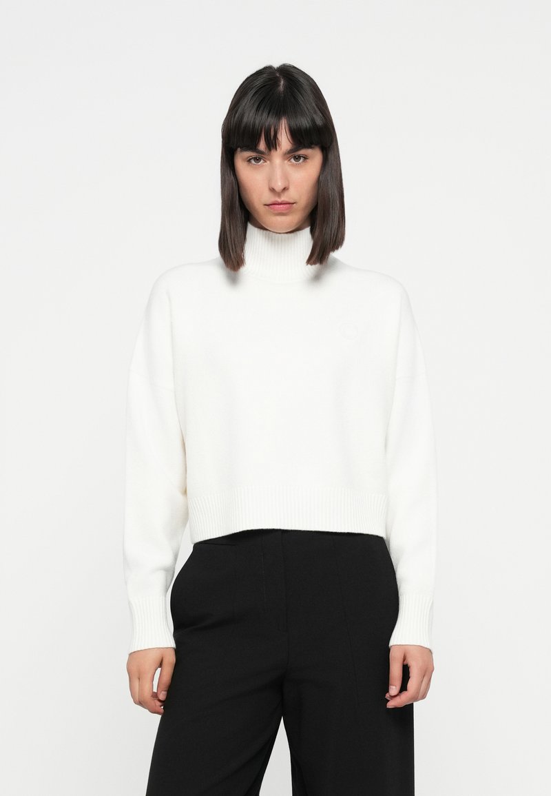 Weißer, taillierter Rollkragenpullover aus weichem Material mit gerippten Bündchen und Saum, getragen über schwarzen High-Waist-Hosen. Keine sichtbaren Muster oder Hardware.