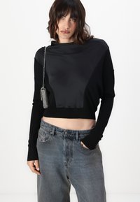 Maglione nero cropped con parte anteriore in satin lucido, abbinato a jeans grigi sbiaditi a vita alta. Tessuto testurizzato e vestibilità rilassata sono elementi notevoli.