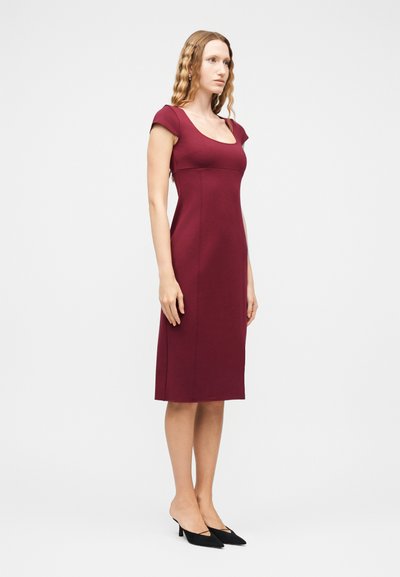 Γυναίκα με κυματιστά μαλλιά, ντυμένη με εφαρμοστό, κοντομάνικο, μπορντό φόρεμα μέχρι το γόνατο και μαύρες γόβες με μυτερή μύτη, στέκεται μπροστά σε λευκό φόντο.