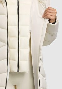 Opblaasjack in off-white met een gewatteerde textuur en een zwarte ritssluiting. Voorzien van een dubbele ritsluiting en een gladde binnenvoering.