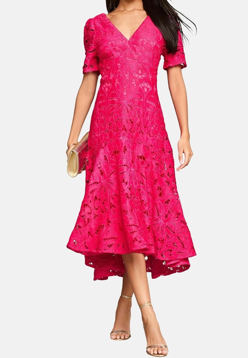 Femme portant une robe en dentelle rose vif à manches courtes et à ourlet évasé, tenant une pochette dorée et portant des talons à bride à la cheville dorés.