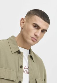 Chemise beige à boutons avec un col, en tissu texturé et deux poches avant. Portée par-dessus une chemise blanche avec du texte brodé en rouge.