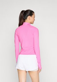 Haut de sport à manches longues rose avec un col haut, texture lisse et accents réfléchissants au dos, associé à des shorts blancs.