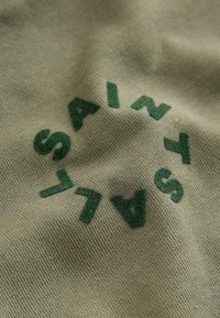 Olivgrüner Stoff mit weicher Textur, der in einem kreisförmigen Layout den grünen Aufdruck "SAINTS" zeigt. Das Design wirkt lässig und modern.