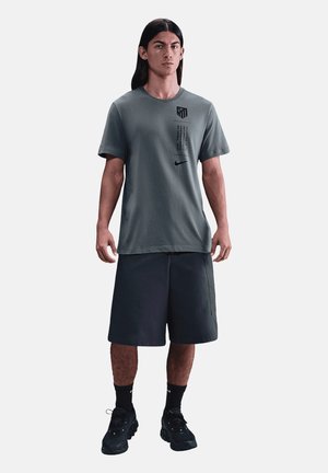 Młody mężczyzna z długimi ciemnymi włosami, ubrany w szarą koszulkę Nike, czarne spodenki, czarne skarpetki Nike i czarne buty sportowe, stoi zwrócony przodem.