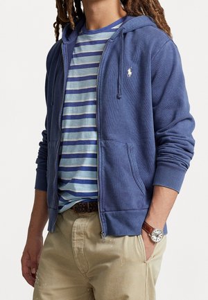 Personne portant un sweat à capuche bleu zippé par-dessus un T-shirt rayé bleu et gris, un pantalon beige et une montre-bracelet avec un bracelet en cuir marron.