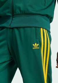 Zeleni atletski trenirka s žutim prugama. Istaknuti detalji uključuju jaknu na zip i izvezeni Adidasov logo. Glatki, poliester materijal.