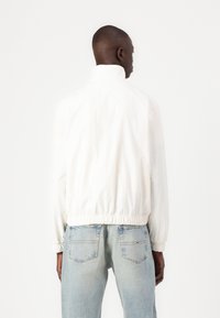 Hvid letvægtsjakke med høj krave, elastisk kant og lange ærmer, parret med lyseblå denimjeans. Glat tekstur, minimalistisk design.