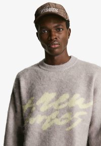 Jasnoszara, dziergana sweter o miękkiej fakturze z żółtym tekstem graficznym. Połączony z brązową, w kratkę czapką baseballową z białym napisem.