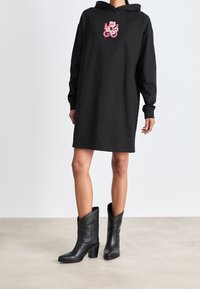 Vestido sudadera negro con detalle de logo floral, mangas largas, corte holgado, que llega a mitad del muslo, combinado con botas negro hasta el tobillo, textura de cuero suave.