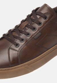 Sneaker in pelle marrone con lacci marroni scuri, suola in gomma e dettagli testurizzati sulla parte superiore. Include un'etichetta del logo vicino ai lacci.