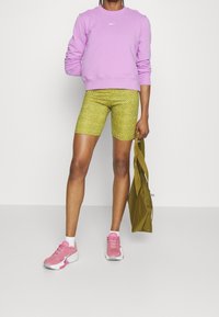 Sweat-shirt violet à col rond, cyclistes jaunes à motifs, et chaussures de sport roses. Un sac vert pend d'une main.