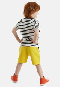 Camiseta a rayas en blanco y negro, pantalones cortos amarillos y zapatillas altas naranjas; un niño está de pie con las manos en las caderas, mirando hacia atrás.