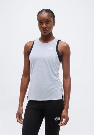 Αθλητική γυναίκα που φοράει αμάνικη ανοιχτό γκρι μπλούζα και μαύρα κολάν με λογότυπα The North Face, ποζάροντας μπροστά σε λευκό φόντο.