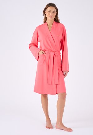 Frau steht barfuß und trägt einen knielangen korallrosa Bademantel mit Gürtel und Fronttaschen, posiert vor einem schlichten hellen Hintergrund.