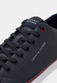 Gros plan sur une sneaker Tommy Hilfiger bleu marine avec des lacets noirs, un œillet rouge, un logo blanc sur le côté et des détails de semelle rouge et blanche.