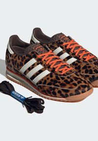 Ζευγάρι αθλητικά παπούτσια Adidas με λεοπάρ σχέδιο, λευκές ρίγες, πορτοκαλί κορδόνια, καφέ σόλες και έξτρα μαύρα κορδόνια δίπλα τους.