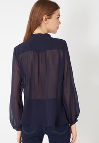 Blusa ligera azul marino, hecha de tejido translúcido, con mangas largas, pliegues en la parte posterior y puños abotonados. Diseño de cuello simple.