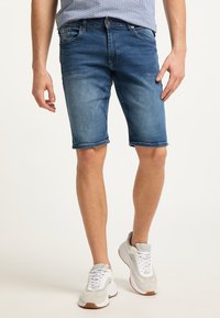 Shorts de mezclilla en lavado azul claro, con un dobladillo desgastado y lazos para cinturón. Combinados con zapatillas deportivas en gris y blanco, que destacan por su diseño deportivo.