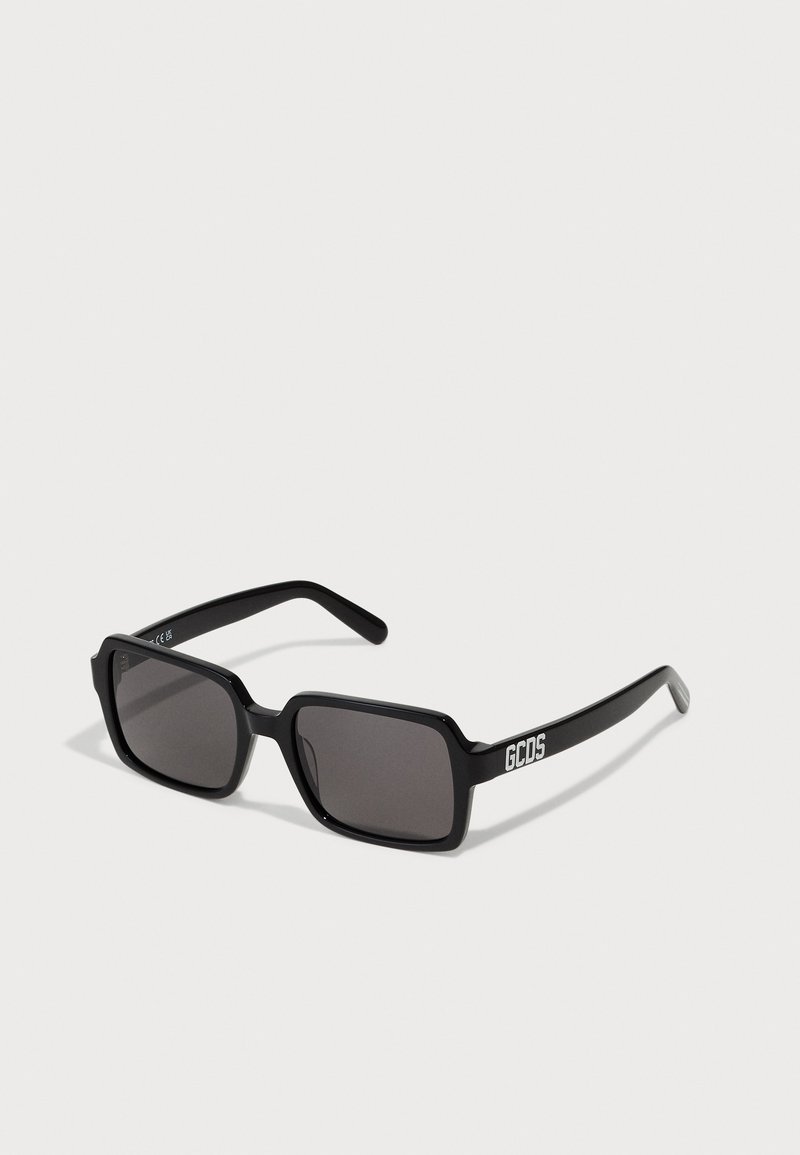 Lunettes de soleil rectangulaires noires avec verres sombres, dotées d'une monture épaisse et de branches avec le logo blanc "GCDS". Surface lisse et design moderne.