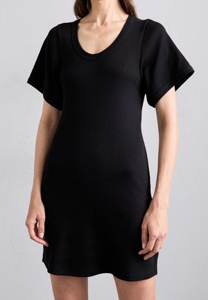 Donna che indossa un vestito nero a maniche corte, a coste, lungo fino al ginocchio con scollatura a barca su sfondo grigio uniforme.