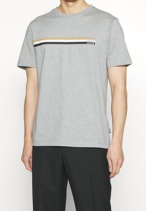 Homme portant un t-shirt gris à manches courtes avec des rayures horizontales beiges, blanches et noires sur la poitrine et le logo "BOSS" sur le côté gauche.
