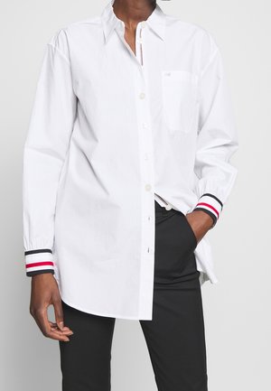 Personne portant une chemise blanche boutonnée avec des poignets rayés rouges, blancs et noirs et un pantalon noir, une main dans la poche, sur un fond uni.