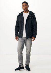 Sort vandtæt jakke med hætte, med lynlåslommer, parret med lysegrå jeans og sorte sneakers med hvide såler.