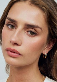 Närbild av ett ansikte med mjuk makeup, slät hy och långa ögonfransar. Bär en guldig hoop-örhänge med en texturerad detalj.