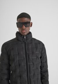 Crna puf jakna s visokim ovratnikom i teksturiranim uzorkom. Ima prednji zipper i bočne džepove, u kombinaciji s oversized crnim naočalama.