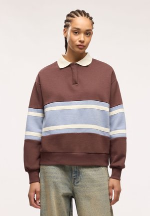 Giovane con capelli intrecciati che indossa una polo a maniche lunghe a righe marroni e blu e jeans chiari, in piedi davanti a uno sfondo semplice.