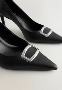 Pantofi negri din piele cu toc înalt și vârfuri ascuțite, având un accesoriu dreptunghiular argintiu pe partea superioară și un finisaj texturat.