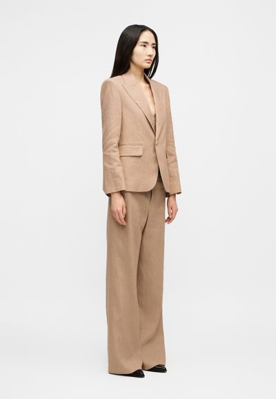 Γυναίκα που στέκεται φορώντας κοστούμι με μπεζ ριγέ σχεδίαση, φαρδιά παντελόνια και σακάκι με ένα κουμπί, κοιτάζοντας μπροστά πάνω σε απλό φόντο.
