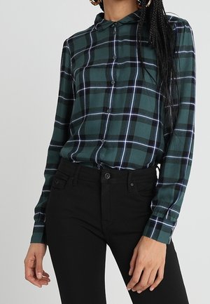 Kobieta ubrana w długą koszulę w zielono-niebieską kratę, wpuszczoną w czarne spodnie z wysokim stanem.