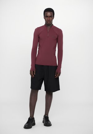 Mørkerød langærmet top med lynlås i halsen, parret med sorte shorts. Outfittet har et stilfuldt, tætsiddende design med en glat tekstur.