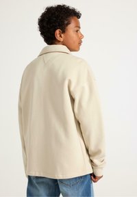 Giacca di cotone beige con colletto, caratterizzata da una vestibilità comoda e cuciture in evidenza. Indossata con jeans in denim blue.