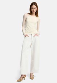 Top beige a maniche lunghe con corpo aderente e maniche testurizzate, abbinato a jeans bianchi a gamba larga. Gli accessori includono scarpe con tacco beige.