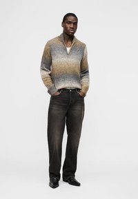Mann trägt einen braun-grauen Strickpullover mit Farbverlauf, schwarze ausgewaschene Jeans und schwarze Schuhe, steht mit den Händen in den Taschen vor einem schlichten Hintergrund.