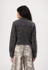 Chaqueta de mezclilla negra recortada con cuello clásico, detalles de costura y puños con botones, combinada con pantalones beige con patrón de serpiente.