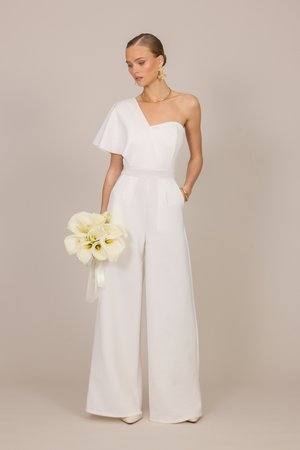 Kvinde i hvid jumpsuit med ét skulderbånd holder en buket hvide calla-liljer, accessoriseret med guldringe, halskæde og armbånd.