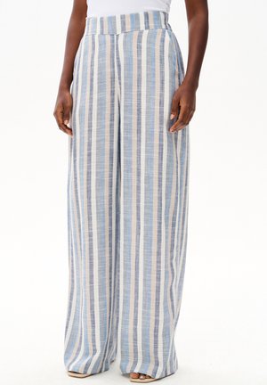Kobieta w spodniach z wysokim stanem i szerokimi nogawkami, z pionowymi niebieskimi, białymi i beżowymi paskami, w połączeniu z sandałami z odkrytymi palcami.