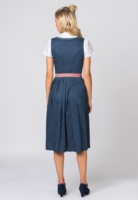 Traditionelles dunkelblaues Kleid mit floralem Muster, plissiertem Rock und einem rosa Bandgürtel. Hat eine weiße Bluse darunter und Absatzschuhe.