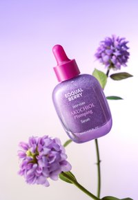 Fioletowa butelka serum z różową zakrętką w kształcie pipety, z błyszczącą teksturą i białym napisem. Na tle delikatnego gradientu i fioletowych kwiatów.